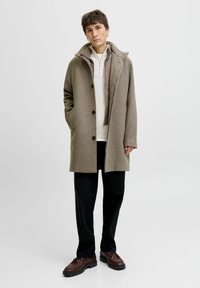 Kahvavillane mantel nuppude sulgemisega, terav krae, küljetaskud, kantud helehallist kattejope ja mustade pükstega; tumepruunid kingad.