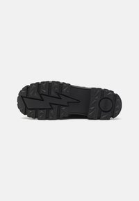Black rubber shoe sole with a zigzag tread pattern, featuring angular spikes for grip. Circular logo on the heel area.