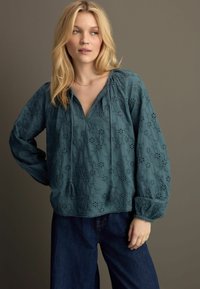 Blusa ricamata color teal con scollo a V, coulisse e maniche lunghe con polsini elastici. Abbinata a pantaloni in denim scuro.