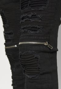 Jeans de mezclilla negra con parches desgastados y roturas. Presenta detalles texturizados y acentos de cremallera plateada en los lados.