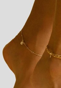 Gul anklet med cirkulære led, små runde perler og en charm med et indgraveret logo, båret om en bar ankel.