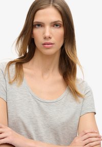 Ung kvinde med lyst brunt hår iført en lysegrå t-shirt, der ser lige frem med armene krydsede foran en ensartet hvid baggrund.