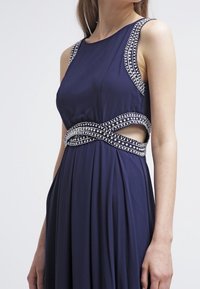 Ärmelloses, marineblaues Kleid mit mit Edelsteinen besetztem Besatz an den Trägern und der Taille, das seitliche Ausschnitte und einen plissierten Rock aufweist.