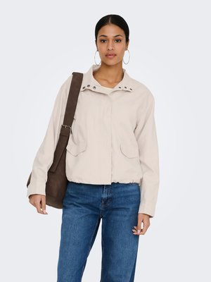 Femme aux cheveux foncés portant une veste beige, un jean bleu, de grandes boucles d’oreilles, portant un sac à épaule marron devant un fond clair uni.