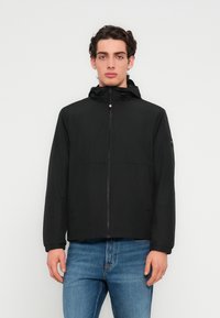 Μαύρο μπουφάν με φερμουάρ και κουκούλα, σχεδιασμένο με απαλό, ελαφρύ ύφασμα και διακριτικές λεπτομέρειες στους ώμους, συνδυασμένο με μπλε τζιν παντελόνι.