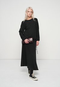 Νεαρή γυναίκα με μακριά ξανθά μαλλιά, φορώντας μια μαύρη μπλούζα, μακριές μαύρες φούστες, μαύρα αθλητικά και κρατώντας μια σκούρα κόκκινη τσάντα.