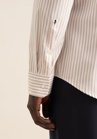 Camicia a strisce beige e bianca, con polsini con bottone e ricamo floreale nero al gomito. Tessuto liscio con vestibilità su misura.