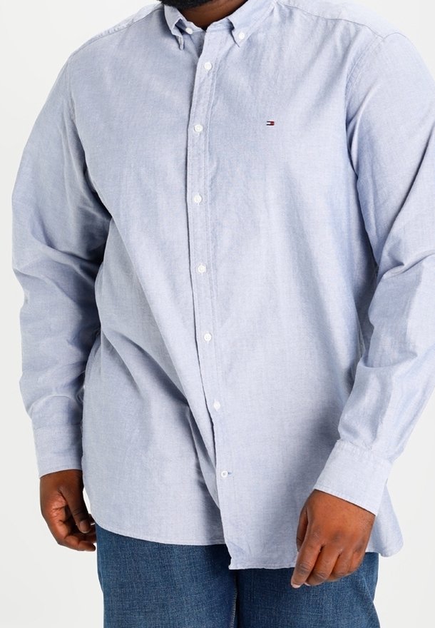 Chemise boutonnée bleu clair en tissu doux, avec un petit logo sur la poitrine, des manches longues et un col classique.