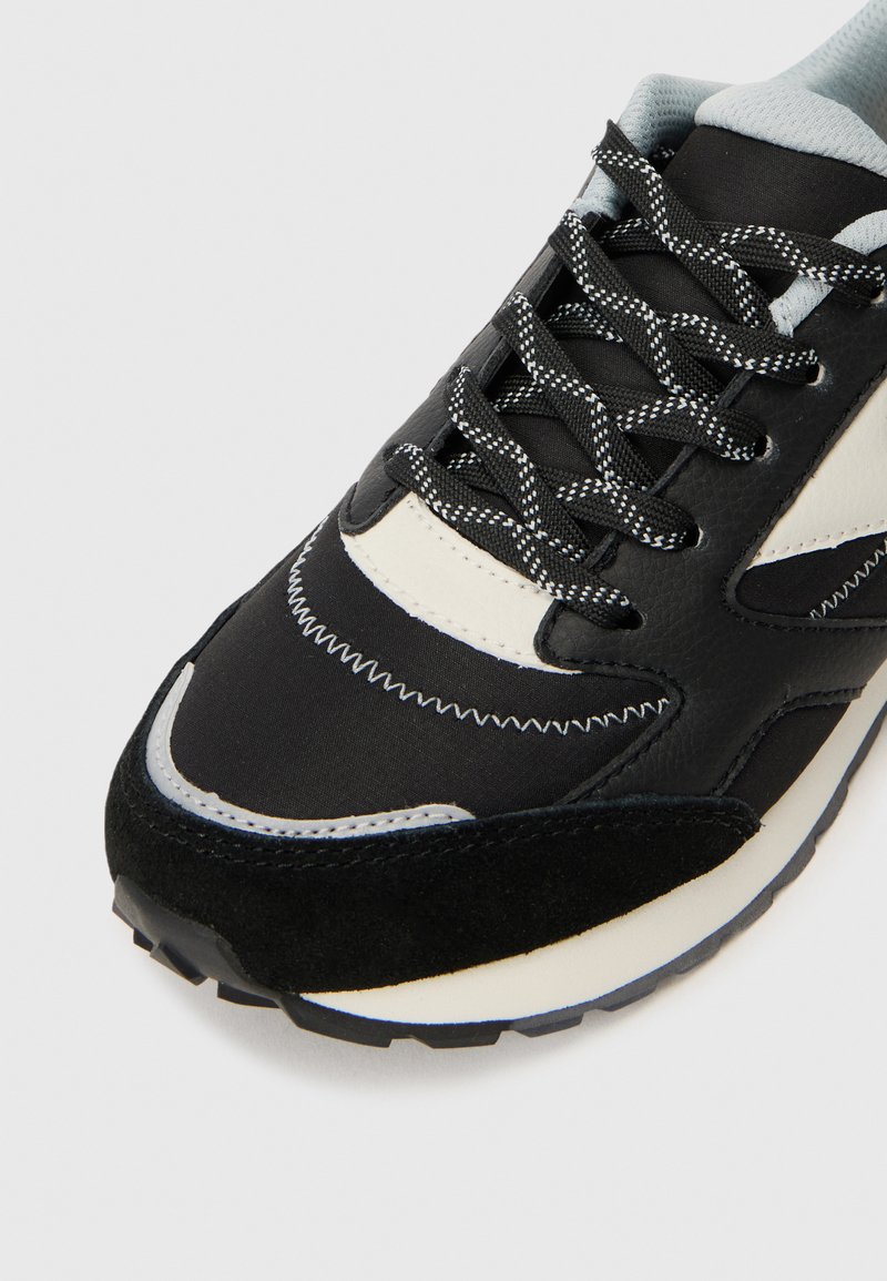 Schwarz-weißer Laufschuh mit gemusterten schwarzen Schnürsenkeln, hellblauem gepolstertem Kragen und weißem Zickzackstich im Bereich der Zehen.