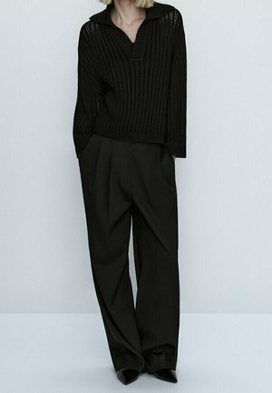 Woman wearing loose black knitted sweater with collar, wide-leg black pants, and black pointed shoes against light background.