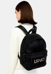 Zaino in nylon nero con forma arrotondata, due spallacci regolabili e una tasca frontale con logo dorato. Tessuto liscio con dettagli minimi.