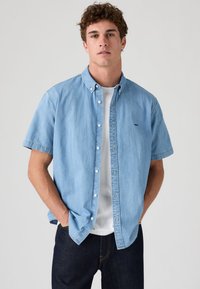 Koszula jeansowa z krótkim rękawem w jasnoniebieskim kolorze z kołnierzem zapinanym na guziki, z widoczną białą podkoszulką pod spodem i ciemnymi dżinsami.