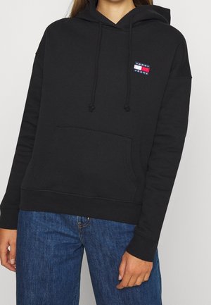 Person iført en sort Tommy Jeans hættetrøje med frontlomme og blå jeans, vist fra hals til hofter mod en ensfarvet baggrund.