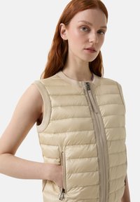 Beige plüschige Weste mit Reißverschluss vorne, geripptem Halsauschnitt und zwei seitlichen Taschen. Mit gestepptem Muster und glatter, glänzender Oberfläche.