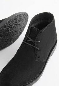 Black suede ankle boots with smooth texture, minimalistic design, and gray laces. Features a textured black rubber sole and stitching accents.