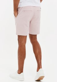 Homme portant un short rose clair, des baskets blanches et une chemise blanche, se tenant les mains dans les poches sur un fond blanc.