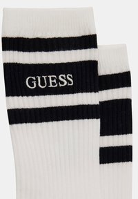 White cotton ribbed socks with black horizontal stripes and "GUESS" logo embroidered in white on the side. Simple athletic design.
