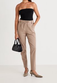 Vrouw in beige hoge taille broek en zwart strapless top, met een zwarte handtas, staand voor een witte achtergrond.