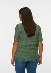 Femme de dos portant un haut en dentelle vert à manches courtes et un jean bleu, avec de longs cheveux bruns, devant un fond uni.