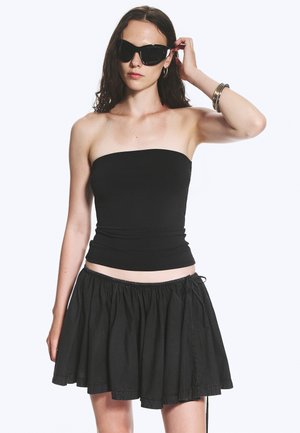 Femme portant un top noir sans bretelles, une jupe noire évasée, de grandes lunettes de soleil noires, des boucles d'oreilles créoles argentées et des bracelets argentés au poignet.