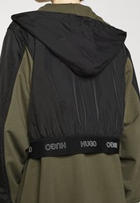 Takki, jossa on musta nylon yläosa ja oliivinvihreä kangas alaosa. Musta elastinen vyötärö, jossa on "HUGO"-logo. Mattapintainen tekstuuri kauttaaltaan.