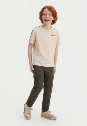 Κοκκινομάλλης αγόρι που φοράει μπεζ μπλούζα με τύπωμα φοινικόδεντρων, ελαιδί παντελόνι καμπαρντίνα και φθαρμένα μπεζ αθλητικά παπούτσια, χαμογελώντας και στέκεται μπροστά σε λευκό φόντο.