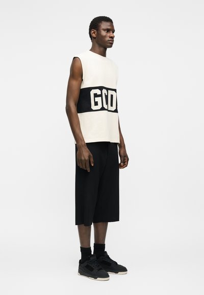 Άνδρας όρθιος φορώντας λευκή αμάνικη μπλούζα με μαύρο κείμενο "GCD", μαύρα σορτς με φαρδύ πόδι, μαύρες κάλτσες και μαύρα αθλητικά παπούτσια πάνω σε λευκό φόντο.