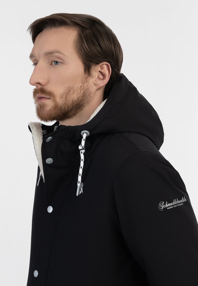 Schwarze Kapuzenjacke mit einem weichen, weißen Fleece-Innenfutter, Druckknopf-Verschluss und kontrastierenden Zugkordeln mit einem gepunkteten Muster. Logo auf dem Ärmel.
