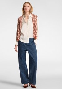 Camicia crema con bottoni e maniche lunghe, abbinata a jeans di denim scuri a gamba larga e a un gilet lavorato a maglia rosso ruggine a fantasia. Scarpe con tacco alte di colore marrone.