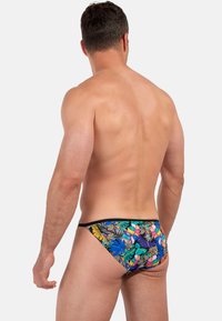 Mann, von hinten gezeigt, trägt farbenfrohe Badeshorts mit tropischem Druck, der Blätter in Blau, Grün, Orange und Lila auf einem weißen Hintergrund zeigt.