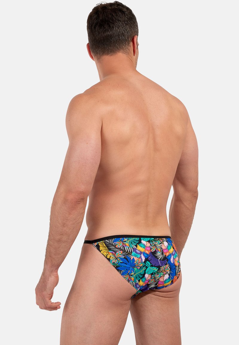 Man gezien van achteren, gekleed in kleurrijke tropische print onderbroek met bladeren in blauw, groen, oranje en paars op een witte achtergrond.