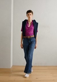 Kortermet rosa skjorte med knapper og et marineblått ermeløst lag, blå bootcut-jeans og hvite sko, satt mot en lys bakgrunn.