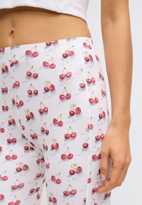 Leggings blancs avec un motif de cerises, présentant des cerises rouges sur de courtes tiges, une texture côtelée et une coupe ajustée. Main posée sur le côté.