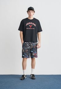 Μαύρο υπερμεγέθες μπλουζάκι με κόκκινο κείμενο "JORDAN" και λογότυπο. Πολύχρωμο σορτς με διάφορα γραφικά μοτίβα. Μαύρο καπέλο και αθλητικά παπούτσια.