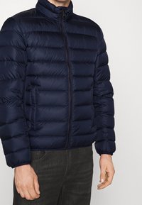 Mörkblå pufferjacka med dragkedja fram, hög krage och vadderad design. Har ett slätt tyg och elastiska ärmmuddar.