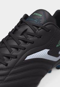 Crampon de soccer en cuir noir avec lacets noirs, bande latérale blanche, logo vert "Joma" sur la languette et crampons bleus visibles sur la semelle.