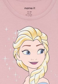 Różowa koszulka z ilustracją postaci z blond włosami, niebieskimi oczami i srebrnymi iskierkami otaczającymi twarz. Okrągły dekolt i miękka tkanina.