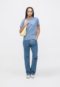 Tricou din bumbac albastru cu detaliu logo, asortat cu blugi rupți de culoare albastru deschis și pantofi bej. Modelul poartă o geantă de umăr galbenă.