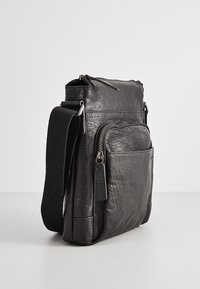 Borsa a tracolla in pelle nera con tasca frontale con zip, superficie testurizzata e tracolla larga in tessuto regolabile, su sfondo semplice.