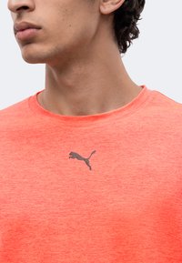 Νεαρό άτομο φοράει κοραλλί ροζ μπλουζάκι Puma, εμφανίζοντας τον λαιμό, τη γνάθο και το λογότυπο Puma στο στήθος σε μονόχρωμο φόντο.