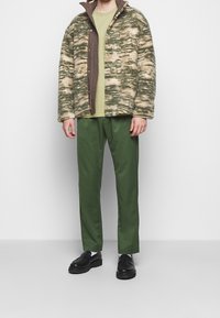 Fleecejacka i beige och grön camouflage mönster med bruna detaljer, tillsammans med en grön t-shirt och gröna byxor, bärs med svarta skor.