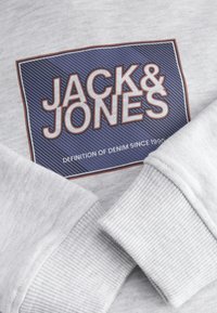 Grå sweatshirt med en blå och röd rektangulär logotyp som har texten "JACK & JONES" med texten "DEFINITION AV DENIM SEDAN 1990" nedanför.