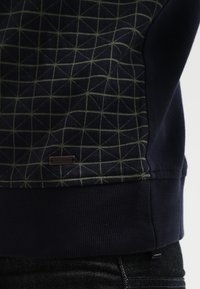 Acercamiento a un suéter azul marino con un patrón de cuadros verdes y una pequeña etiqueta de metal de la marca cerca del dobladillo acanalado, usado sobre jeans oscuros.