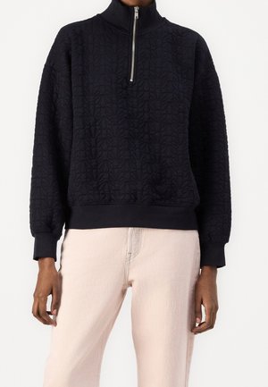 Czarny pikowany pulower z kwiatowym wzorem i przednim zamkiem błyskawicznym, noszony z jasnobeżowymi spodniami, widoczny od szyi do bioder.