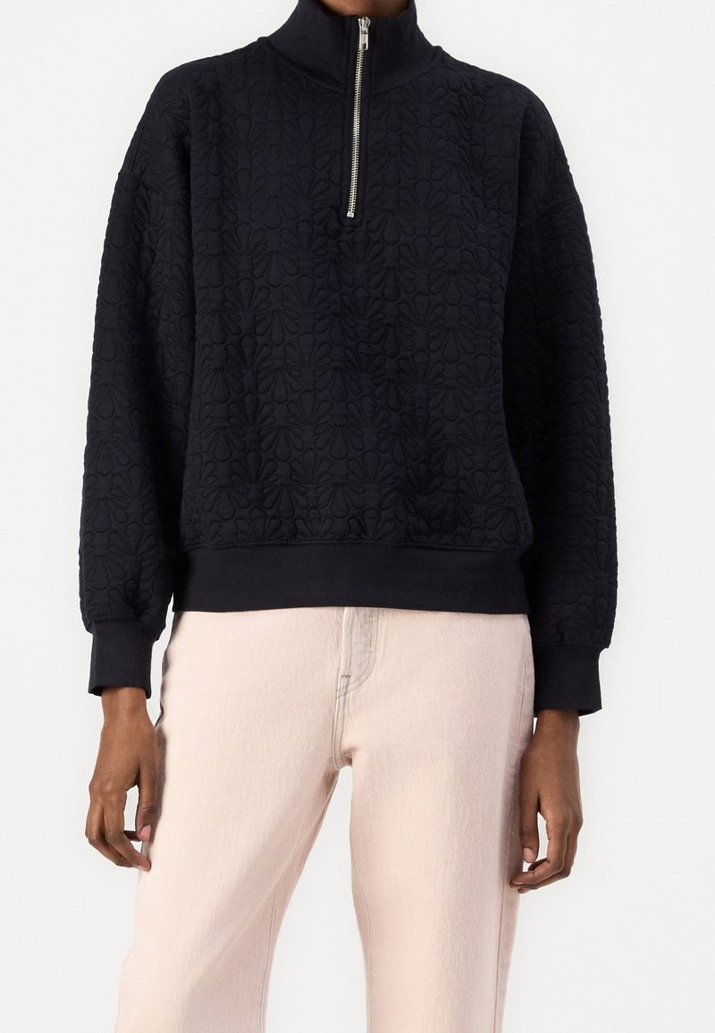 Pull noir matelassé avec motif floral et fermeture éclair à l'avant, porté avec un pantalon beige clair, visible du cou aux hanches.