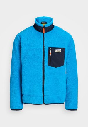 Niebieska polarowa kurtka z granatową kieszenią na piersi, zamek błyskawiczny, wysoki kołnierz i elastyczne mankiety. Teksturowana tkanina z wyrazistymi kontrastami kolorystycznymi.
