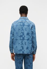 Άνδρας που φορά μπλε τζιν μπουφάν και παντελόνι με έντονο αφηρημένο λουλουδάτο μοτίβο, στέκεται με την πλάτη προς τα εμπρός σε απλό λευκό φόντο.