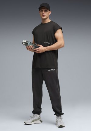 Camiseta sin mangas negra con texto blanco, pantalones jogger grises con logo blanco, zapatillas deportivas gris claro, sosteniendo una botella de agua transparente.