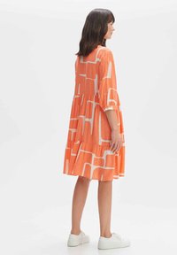 Orangenes Kleid mit weißen abstrakten Mustern, das lange Ärmel, ein gestuftes Design und einen lässigen Schnitt aufweist, kombiniert mit weißen Sneakers.