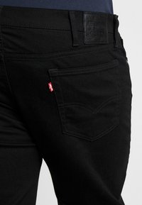 Černé džíny z denimu s tradičním designem pěti kapes, s koženým štítkem na zadní straně a červenou tabulkou na zadní kapse.